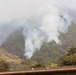 A wildfire burns on a heavily vegetated hillside, producing large plumes of white-gray smoke that obscure the upper portion of the mountain. In the foreground, three firefighters wearing yellow protective gear and hard hats are visible standing on the roof of a building, apparently monitoring the fire situation. The scene suggests an active wildfire response with structures potentially at risk.