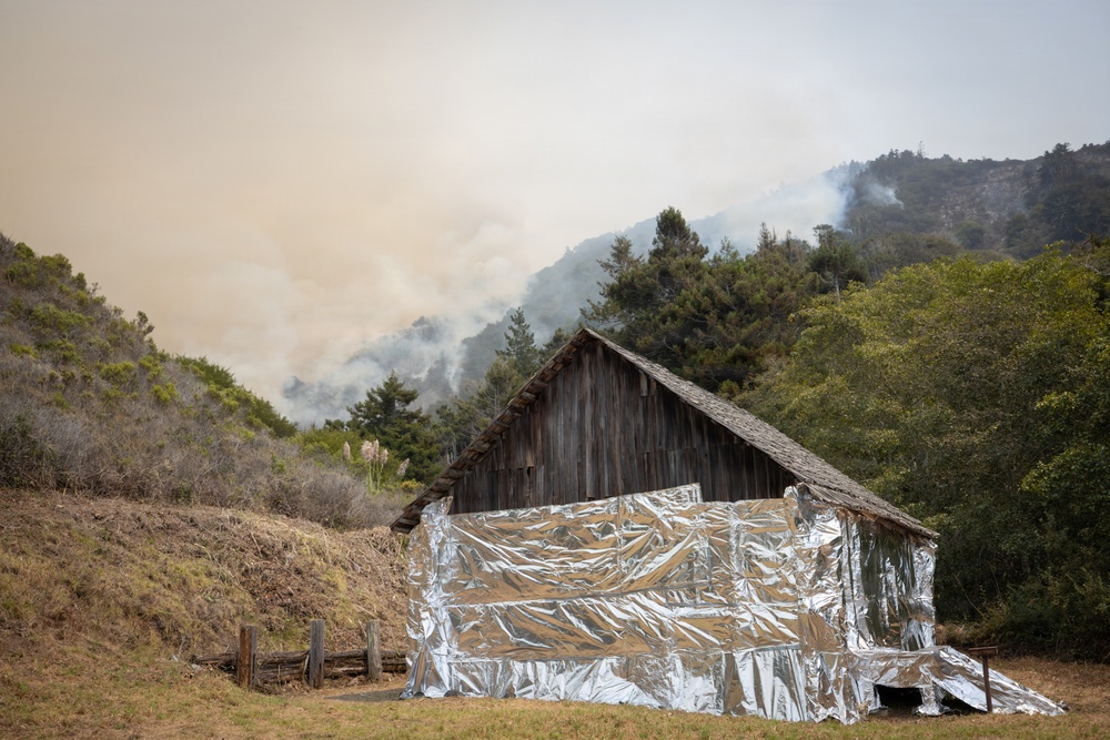 A rustic wooden barn has been wrapped with reflective emergency fire-resistant foil sheeting on its exterior walls as a wildfire protection measure. In the background, active wildfire smoke is visible rising from a hillside covered with trees and vegetation. The sky is heavily hazy and discolored with smoke, indicating an active and nearby wildfire event.