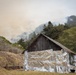 A rustic wooden barn has been wrapped with reflective emergency fire-resistant foil sheeting on its exterior walls as a wildfire protection measure. In the background, active wildfire smoke is visible rising from a hillside covered with trees and vegetation. The sky is heavily hazy and discolored with smoke, indicating an active and nearby wildfire event.