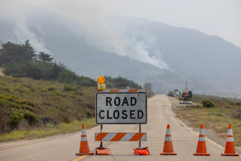 A road closure barricade with a 'ROAD CLOSED' sign and orange traffic cones blocks a two-lane highway. In the background, smoke rises from an active wildfire burning on a hillside. Emergency or utility vehicles are visible further down the road near the fire zone.
