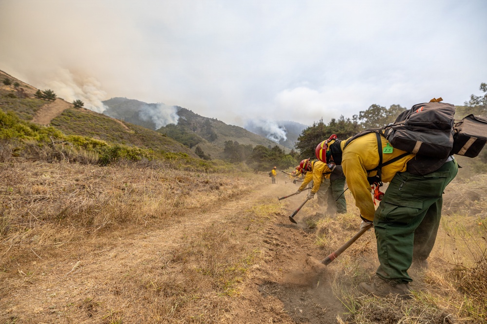 A crew of wildland firefighters in yellow Nomex protective gear and red helmets are constructing a firebreak by scraping a dirt line along a hillside trail. Smoke is visible rising from a fire burning on the hillsides in the background. The crew appears to be working in a line formation using hand tools such as Pulaskis and McLeods to clear vegetation and expose bare soil.