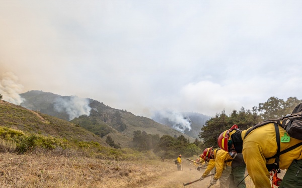 A crew of wildland firefighters in yellow Nomex protective gear and red helmets are constructing a firebreak by scraping a dirt line along a hillside trail. Smoke is visible rising from a fire burning on the hillsides in the background. The crew appears to be working in a line formation using hand tools such as Pulaskis and McLeods to clear vegetation and expose bare soil.