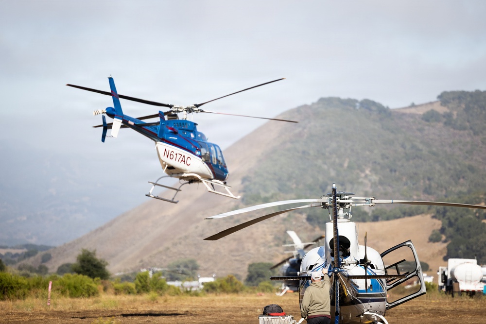 A blue and white helicopter with tail number N617AC is captured in a low hover or takeoff above a grassy field, with mountainous terrain visible in the background. In the foreground, a second grounded helicopter is visible with a person standing nearby, apparently performing ground crew duties. The scene appears to be a temporary helicopter operations area or staging ground.