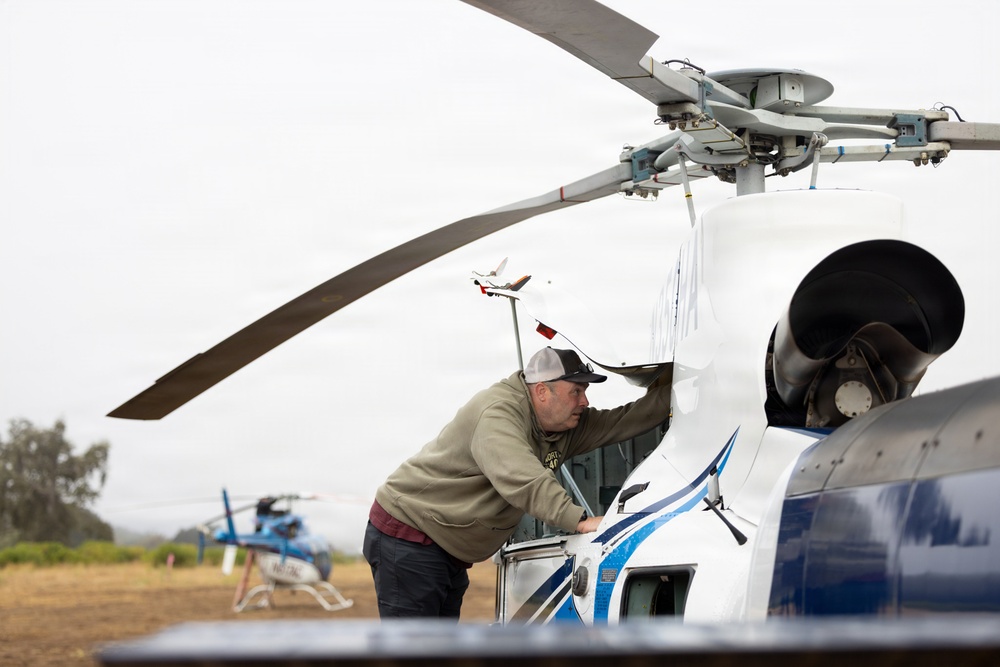 A man in a baseball cap and sweatshirt is inspecting or performing maintenance on a large white helicopter with blue stripe markings. The scene appears to be at an open field or temporary landing area, with a second smaller blue helicopter visible in the background. The image captures a ground-level maintenance or pre-flight check operation.