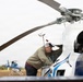 A man in a baseball cap and sweatshirt is inspecting or performing maintenance on a large white helicopter with blue stripe markings. The scene appears to be at an open field or temporary landing area, with a second smaller blue helicopter visible in the background. The image captures a ground-level maintenance or pre-flight check operation.
