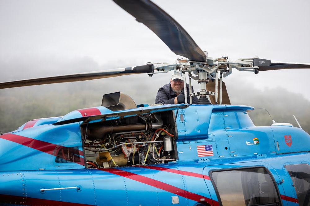 A mechanic or technician is performing maintenance on a blue and red helicopter with an open engine compartment. The man is leaning over the top of the aircraft near the main rotor assembly, inspecting or working on the helicopter. The aircraft displays an American flag decal and what appears to be company branding on its fuselage.