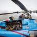 A mechanic or technician is performing maintenance on a blue and red helicopter with an open engine compartment. The man is leaning over the top of the aircraft near the main rotor assembly, inspecting or working on the helicopter. The aircraft displays an American flag decal and what appears to be company branding on its fuselage.
