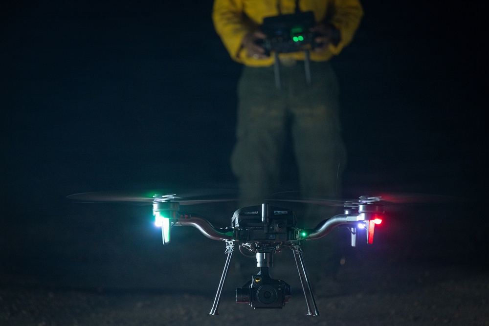 A quadcopter drone is captured hovering at low altitude during nighttime operations, equipped with a gimbal-mounted camera and illuminated navigation lights. A drone operator wearing a yellow jacket and olive/green pants stands in the background holding a remote controller. The image is taken with a shallow depth of field, keeping the drone in sharp focus while the operator remains blurred.