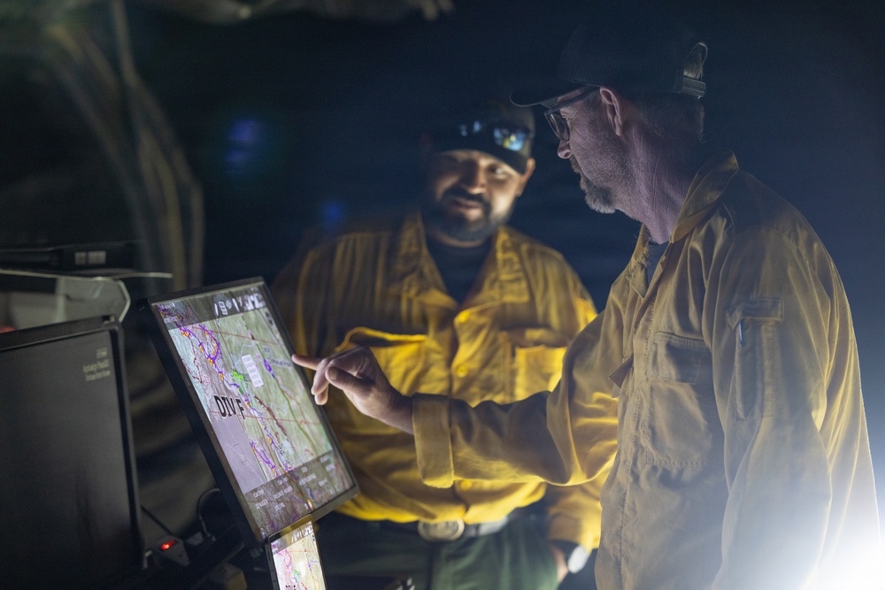Two individuals wearing yellow fire-resistant shirts and caps are reviewing a digital mapping display at night. One person is pointing at the screen, which appears to show a topographical or incident command map with the label 'DIV F' visible. The setting suggests a wildfire operations command post or field briefing.