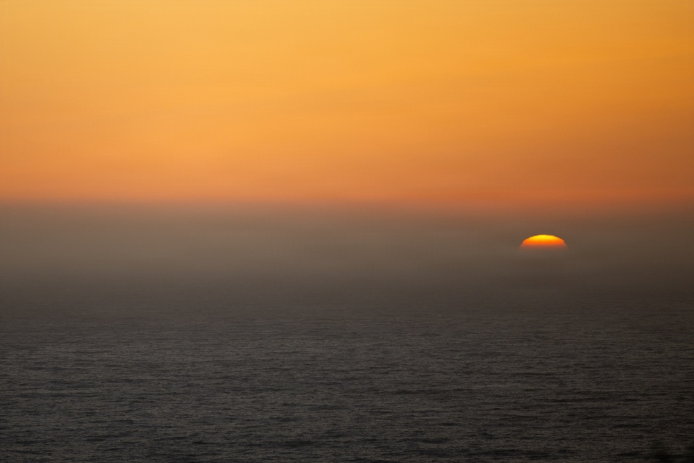 A coastal sunset scene showing the sun partially setting below the horizon over an open ocean. A distinct horizontal band of sea fog or marine layer obscures the middle portion of the scene, with the sun visible just above the water line on the right side. The sky is dominated by warm orange and amber tones.