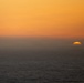 A coastal sunset scene showing the sun partially setting below the horizon over an open ocean. A distinct horizontal band of sea fog or marine layer obscures the middle portion of the scene, with the sun visible just above the water line on the right side. The sky is dominated by warm orange and amber tones.