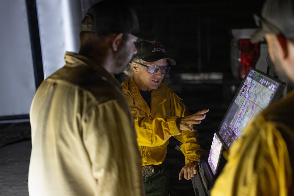 Three individuals wearing yellow Nomex fire-resistant shirts and baseball caps are gathered around computer monitors displaying what appears to be a topographic or fire mapping system. The central figure, a woman wearing blue-tinted glasses and a cap with 'Cleveland National Forest' text, is actively gesturing toward the screens and appears to be briefing or directing the others. The setting appears to be a mobile command post or incident command facility.