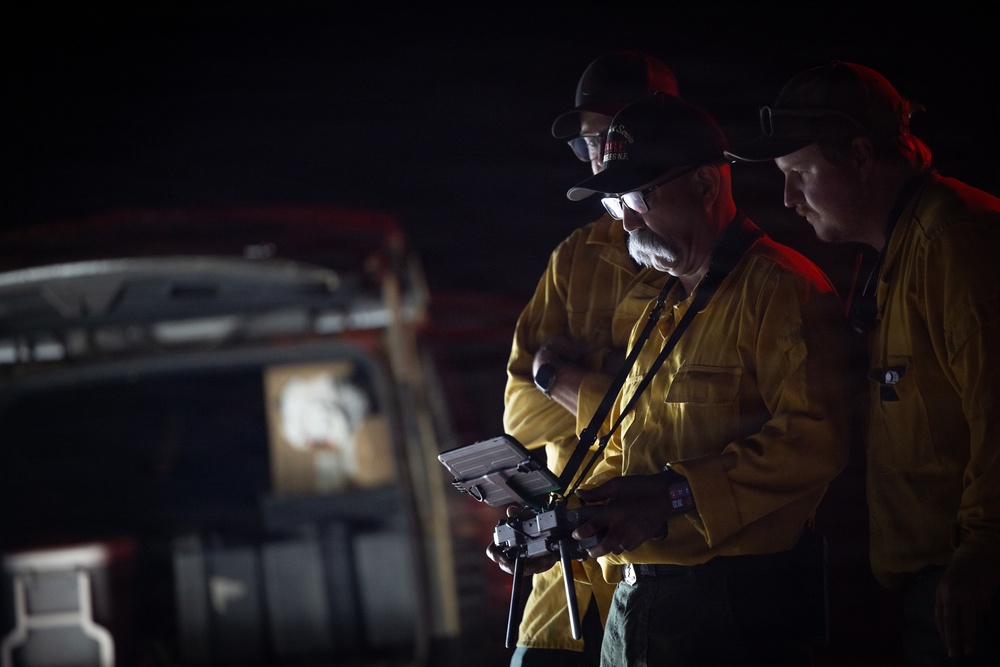 Two individuals wearing yellow fire-resistant shirts and black caps are operating a drone controller with a tablet attachment during nighttime conditions. The scene appears to be an emergency or wildfire response operation, with a vehicle visible in the dark background. The primary operator is illuminated by the screen light while reviewing data on the mounted tablet display.