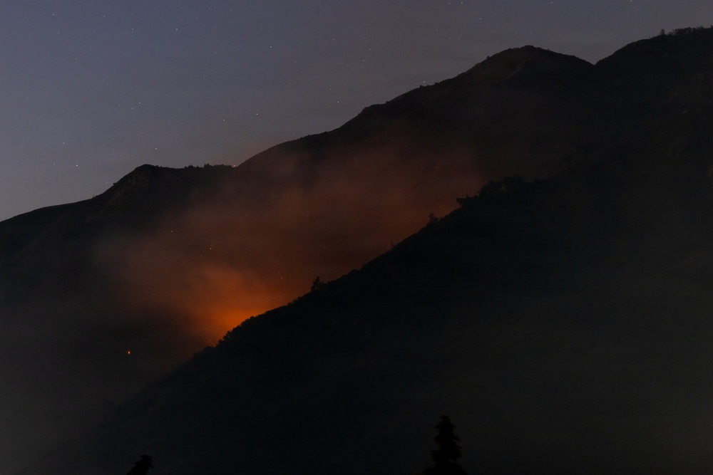 A nighttime photograph capturing an active wildfire burning on a mountainside. Intense orange and red flames illuminate smoke billowing across the dark mountain slope. Stars are faintly visible in the twilight sky above the silhouetted mountain ridgeline.