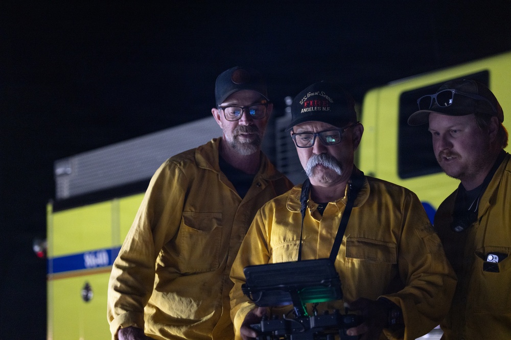 Three men wearing yellow fire-resistant shirts are gathered at night near a yellow emergency vehicle. The central figure, wearing a cap reading 'U.S. Forest Service Fire Angeles N.F.', is holding what appears to be a drone controller or electronic device. They appear to be monitoring or operating equipment during a nighttime fire response operation.
