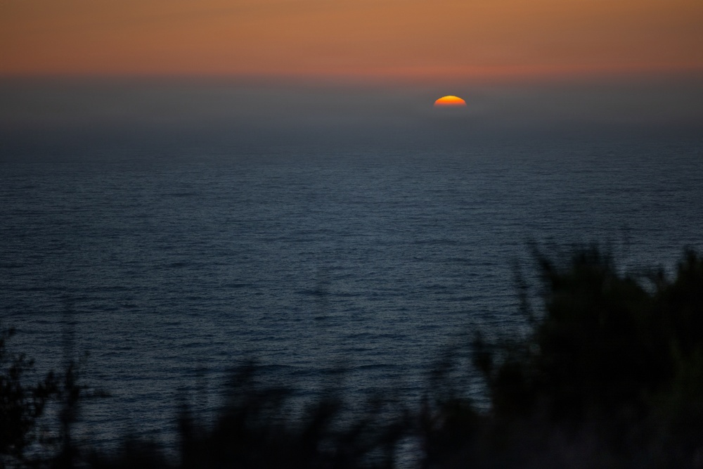 A coastal sunset photograph capturing the final moments of the sun as it dips below the ocean horizon. Only a small semicircle of deep orange sun remains visible above the waterline, set against a darkening blue-grey sea and warm amber sky. Silhouetted vegetation is visible in the foreground lower portion of the frame.