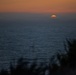 A coastal sunset photograph capturing the final moments of the sun as it dips below the ocean horizon. Only a small semicircle of deep orange sun remains visible above the waterline, set against a darkening blue-grey sea and warm amber sky. Silhouetted vegetation is visible in the foreground lower portion of the frame.