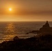 A coastal sunset scene captured from an elevated vantage point overlooking the ocean. The sun sits low on the horizon, casting a warm golden-orange glow across the water and sky, with a prominent rocky outcropping silhouetted in the foreground. A small tree or shrub is visible atop the largest rock formation, adding a distinctive detail to the silhouette.
