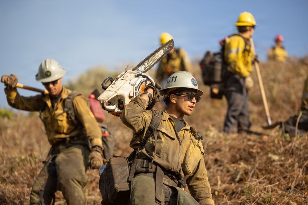 A crew of wildland firefighters is working on a hillside, carrying tools and equipment. The foreground shows a firefighter carrying a chainsaw on their shoulder, while others carry hand tools such as axes and shovels. The crew is wearing standard wildland firefighting gear including Nomex suits, hard hats, and protective equipment.