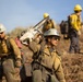 A crew of wildland firefighters is working on a hillside, carrying tools and equipment. The foreground shows a firefighter carrying a chainsaw on their shoulder, while others carry hand tools such as axes and shovels. The crew is wearing standard wildland firefighting gear including Nomex suits, hard hats, and protective equipment.