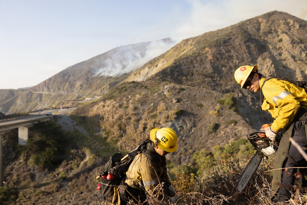 Two wildland firefighters in yellow protective gear are working on a steep hillside with an active wildfire visible in the background producing smoke on the mountain ridge. A highway overpass or bridge is visible in the valley below. The firefighters appear to be conducting vegetation clearing or fire line operations using a chainsaw.