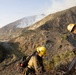 Two wildland firefighters in yellow protective gear are working on a steep hillside with an active wildfire visible in the background producing smoke on the mountain ridge. A highway overpass or bridge is visible in the valley below. The firefighters appear to be conducting vegetation clearing or fire line operations using a chainsaw.