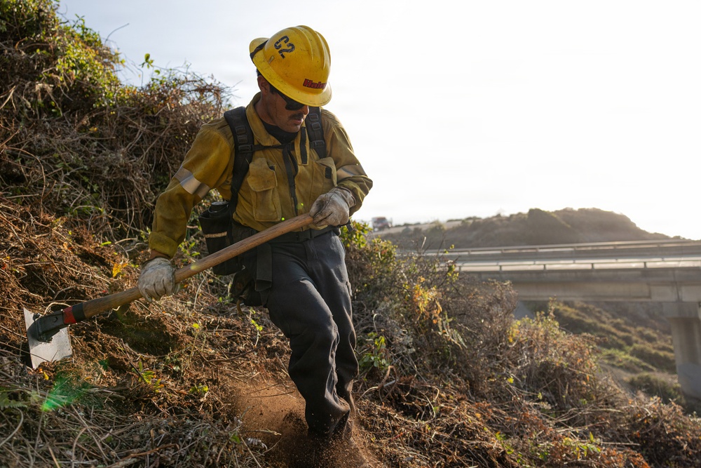 A wildland firefighter wearing standard yellow Nomex fire-resistant gear and a yellow hard hat marked 'C2' is using a Pulaski axe-hoe tool to cut a firebreak on a steep, dry hillside. The worker appears to be part of a hand crew performing line construction or mop-up operations near a highway overpass or bridge visible in the background. The terrain is covered in dry, dead brush consistent with fire-prone chaparral vegetation.