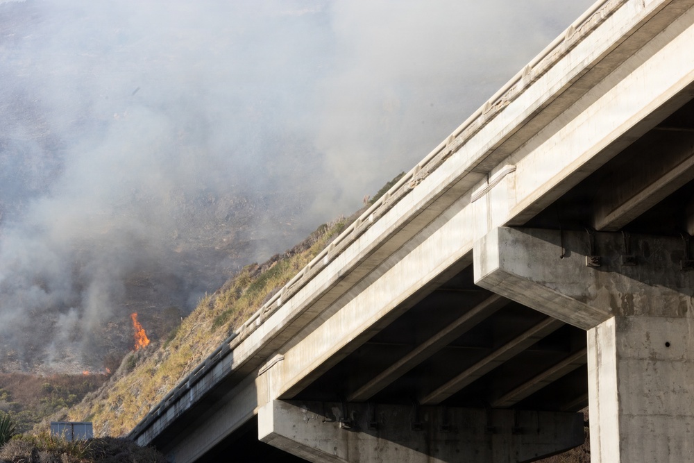 A concrete overpass or highway bridge is photographed from below at an angle, with an active wildfire burning on a hillside visible in the background. Thick gray-white smoke fills the upper portion of the image, and open flames are visible on the vegetation-covered slope. The fire appears to be in close proximity to the infrastructure.