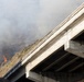 A concrete overpass or highway bridge is photographed from below at an angle, with an active wildfire burning on a hillside visible in the background. Thick gray-white smoke fills the upper portion of the image, and open flames are visible on the vegetation-covered slope. The fire appears to be in close proximity to the infrastructure.