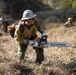 A wildland firefighter wearing a white helmet marked 'C11' carries a STIHL chainsaw while moving across dry, grassy terrain. The worker is equipped with standard wildland firefighting gear including a yellow Nomex shirt, green pants, gloves, and a backpack with a fuel canister. Additional crew members are visible in the background, suggesting a coordinated fire suppression or fuel reduction operation.