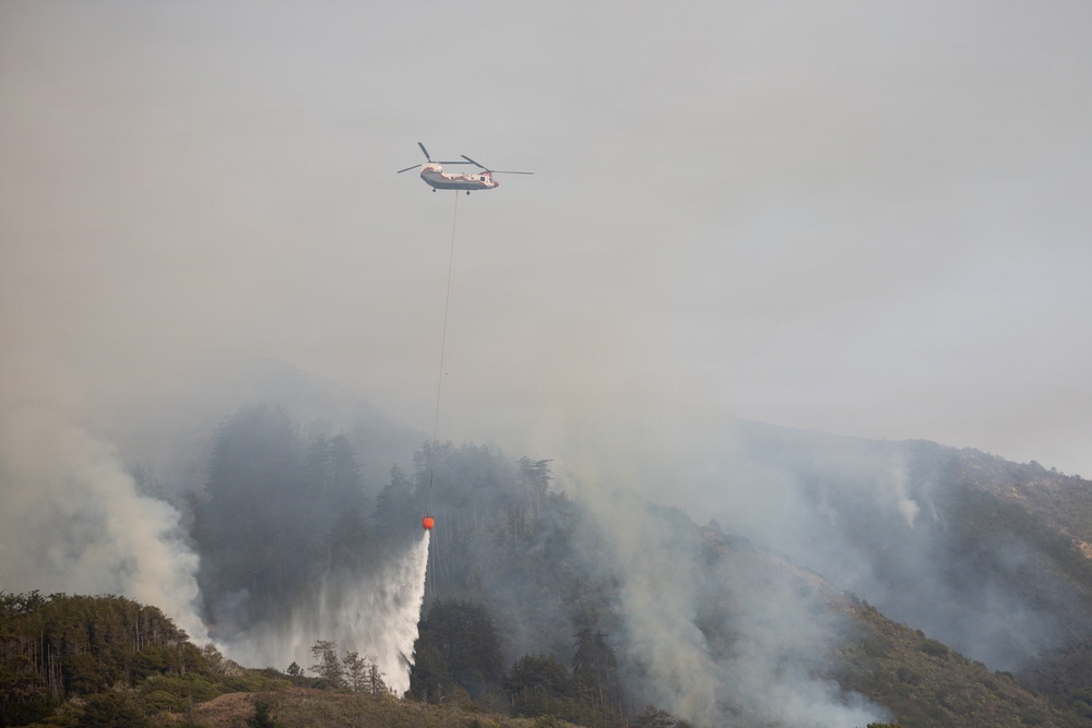 A large tandem-rotor helicopter (consistent with a Boeing CH-47 Chinook or similar heavy-lift aircraft) is conducting aerial firefighting operations over a forested hillside. The helicopter is deploying water from an orange Bambi bucket suspended by a long line, releasing a large cascade of water onto the burning vegetation below. The scene is heavily obscured by thick white and gray smoke rising from multiple active fire areas across the hillside.