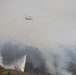 A large tandem-rotor helicopter (consistent with a Boeing CH-47 Chinook or similar heavy-lift aircraft) is conducting aerial firefighting operations over a forested hillside. The helicopter is deploying water from an orange Bambi bucket suspended by a long line, releasing a large cascade of water onto the burning vegetation below. The scene is heavily obscured by thick white and gray smoke rising from multiple active fire areas across the hillside.