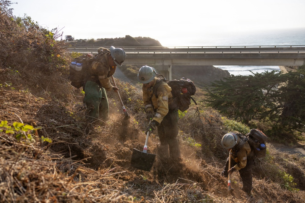 Three wildland firefighters in full protective gear are working on a dry, brush-covered hillside slope, digging and clearing vegetation likely to create a firebreak. The scene is set along a coastal highway, with a bridge, ocean, and rocky headland visible in the background. The workers are equipped with hand tools including shovels and appear to be part of a hotshot or hand crew operation.