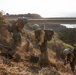 Three wildland firefighters in full protective gear are working on a dry, brush-covered hillside slope, digging and clearing vegetation likely to create a firebreak. The scene is set along a coastal highway, with a bridge, ocean, and rocky headland visible in the background. The workers are equipped with hand tools including shovels and appear to be part of a hotshot or hand crew operation.