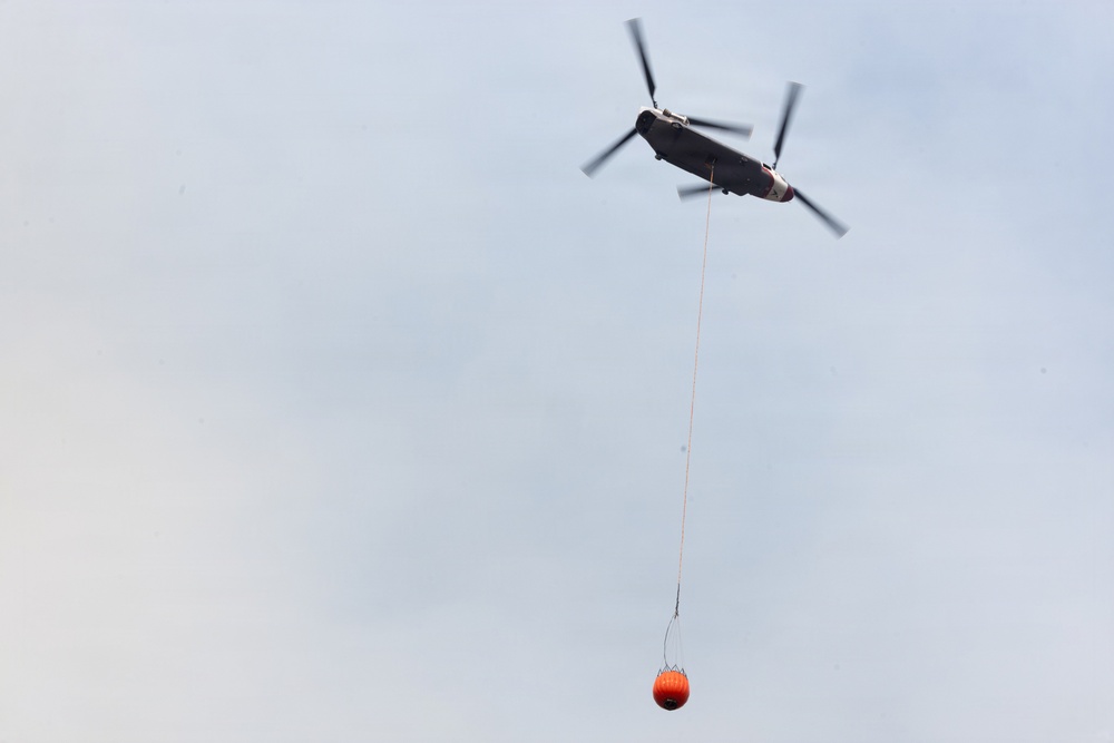A large tandem-rotor helicopter, consistent with a Boeing CH-47 Chinook configuration, is flying through an overcast sky while carrying an orange firefighting water bucket suspended on a long line beneath it. The helicopter appears to be engaged in aerial firefighting operations, transporting the bambi bucket likely to or from a water source. The image is taken from a low angle looking upward.