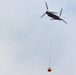 A large tandem-rotor helicopter, consistent with a Boeing CH-47 Chinook configuration, is flying through an overcast sky while carrying an orange firefighting water bucket suspended on a long line beneath it. The helicopter appears to be engaged in aerial firefighting operations, transporting the bambi bucket likely to or from a water source. The image is taken from a low angle looking upward.