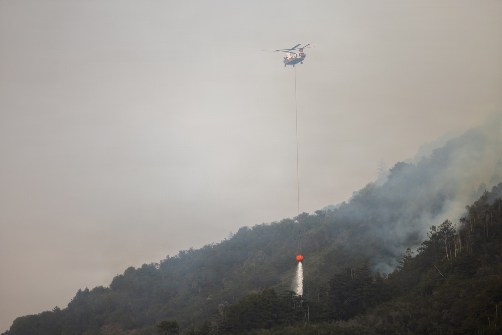 A firefighting helicopter is conducting aerial water-dropping operations over a forested hillside engulfed in a wildfire. The helicopter is carrying an orange bambi bucket suspended by a long cable, actively releasing water onto the burning vegetation below. Heavy smoke fills the atmosphere, significantly reducing visibility across the scene.