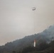 A firefighting helicopter is conducting aerial water-dropping operations over a forested hillside engulfed in a wildfire. The helicopter is carrying an orange bambi bucket suspended by a long cable, actively releasing water onto the burning vegetation below. Heavy smoke fills the atmosphere, significantly reducing visibility across the scene.