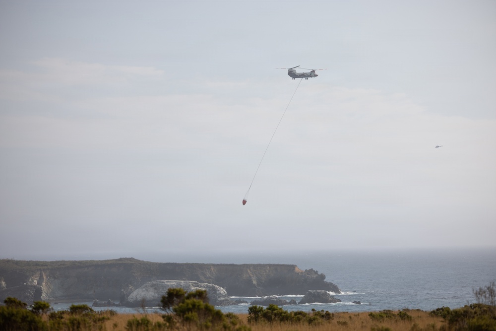 A tandem-rotor helicopter, consistent with a CH-47 Chinook type, is flying over a rugged coastal landscape while carrying a suspended firefighting water bucket (helibucket) on a long line. The scene appears to depict aerial firefighting or water-scooping operations near a rocky coastline. A second, smaller aircraft is faintly visible in the background to the right.