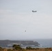 A tandem-rotor helicopter, consistent with a CH-47 Chinook type, is flying over a rugged coastal landscape while carrying a suspended firefighting water bucket (helibucket) on a long line. The scene appears to depict aerial firefighting or water-scooping operations near a rocky coastline. A second, smaller aircraft is faintly visible in the background to the right.