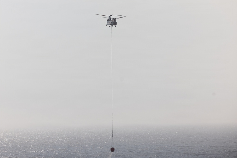 A tandem-rotor helicopter, consistent with a CH-47 Chinook design, is flying over a body of water while suspending a firefighting water bucket on a long cable. The bucket appears to be at or near the water surface, suggesting the helicopter is in the process of filling it. The scene is captured in hazy, overcast conditions with muted tones.