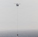A tandem-rotor helicopter, consistent with a CH-47 Chinook design, is flying over a body of water while suspending a firefighting water bucket on a long cable. The bucket appears to be at or near the water surface, suggesting the helicopter is in the process of filling it. The scene is captured in hazy, overcast conditions with muted tones.
