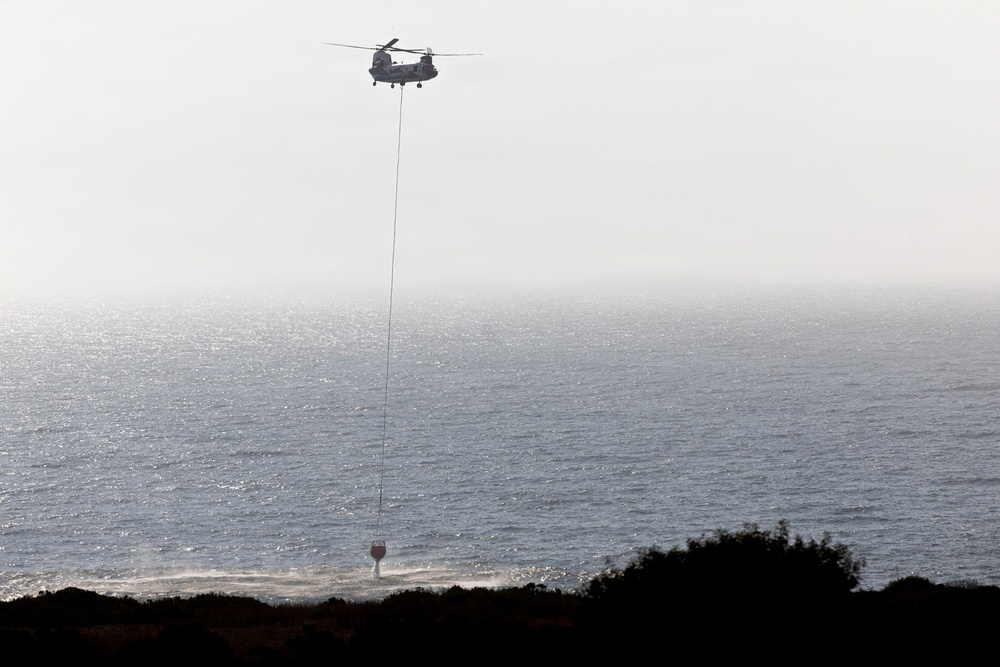 A tandem-rotor helicopter, consistent with a CH-47 Chinook design, is hovering over a coastal area while suspending a water bucket on a long line. The bucket appears to be dipping into or near the water's surface, suggesting a water-scooping operation likely associated with aerial firefighting. The scene is set against a hazy ocean backdrop with silhouetted coastal vegetation in the foreground.