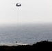 A tandem-rotor helicopter, consistent with a CH-47 Chinook design, is hovering over a coastal area while suspending a water bucket on a long line. The bucket appears to be dipping into or near the water's surface, suggesting a water-scooping operation likely associated with aerial firefighting. The scene is set against a hazy ocean backdrop with silhouetted coastal vegetation in the foreground.