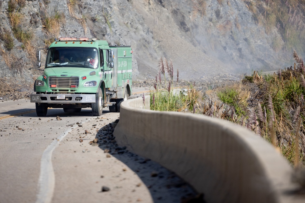 A green California fire department truck (marked CA-LPF E574) is traveling along a narrow mountain road that has debris scattered across it, suggesting a recent rockslide or landslide. The road runs alongside a steep rocky cliff face with smoke or dust visible in the background. The scene indicates an active emergency response situation in a rugged coastal or mountain terrain.