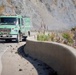 A green California fire department truck (marked CA-LPF E574) is traveling along a narrow mountain road that has debris scattered across it, suggesting a recent rockslide or landslide. The road runs alongside a steep rocky cliff face with smoke or dust visible in the background. The scene indicates an active emergency response situation in a rugged coastal or mountain terrain.