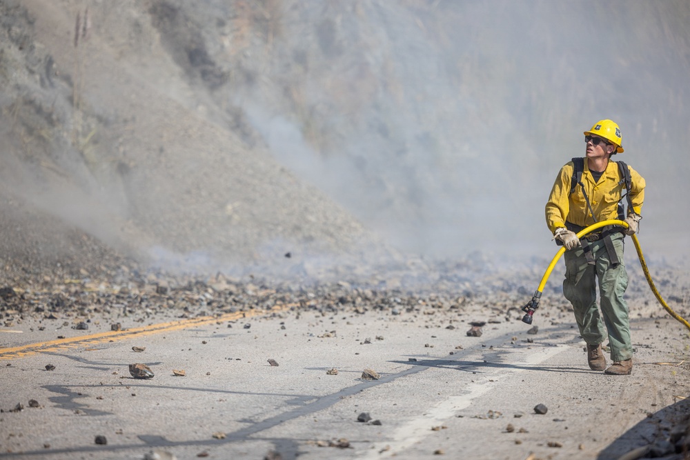 A wildland firefighter in standard yellow Nomex gear is working on a smoke-filled road covered in debris, likely near a hillside or cliff face. The firefighter is actively pulling or advancing a yellow fire hose toward a smoke source. The scene suggests an active wildfire or post-rockslide fire situation along a paved two-lane road.