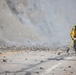 A wildland firefighter in standard yellow Nomex gear is working on a smoke-filled road covered in debris, likely near a hillside or cliff face. The firefighter is actively pulling or advancing a yellow fire hose toward a smoke source. The scene suggests an active wildfire or post-rockslide fire situation along a paved two-lane road.