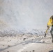 A wildland firefighter in standard protective gear is running along a paved road while carrying a yellow fire hose. The scene is surrounded by smoke, scorched earth, and debris, indicating an active wildfire operation. The firefighter appears to be repositioning the hose line in response to fire conditions.