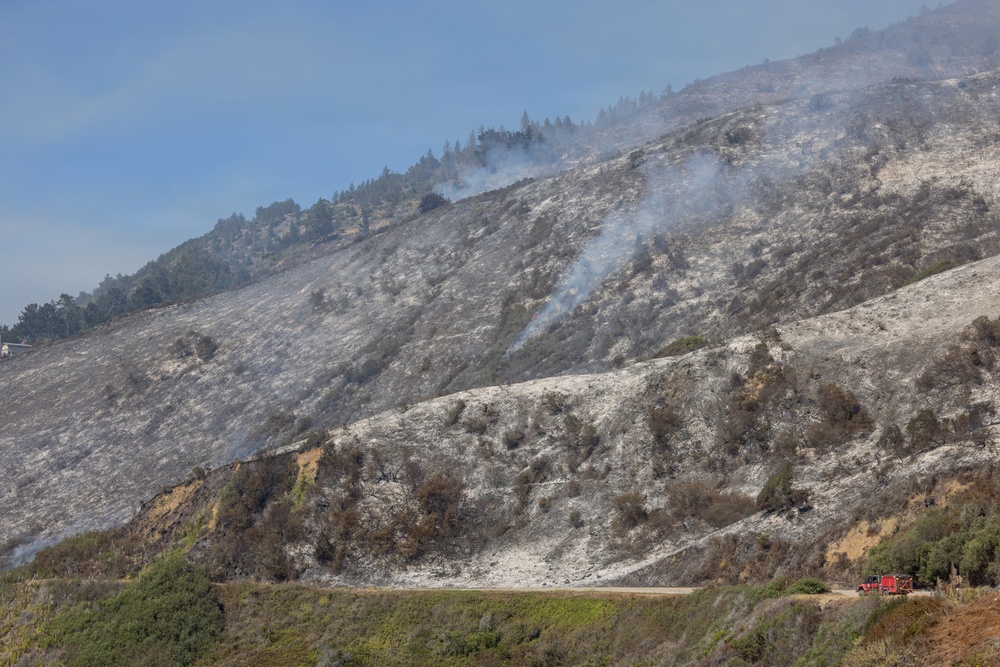 A large hillside or mountain slope shows extensive wildfire burn damage, with the majority of vegetation reduced to ash and charred remnants. Multiple columns of smoke are still rising from smoldering areas across the slope, indicating the fire is recently extinguished or still partially active. A red fire truck is visible on a road at the lower right, suggesting active firefighting or post-fire monitoring operations.